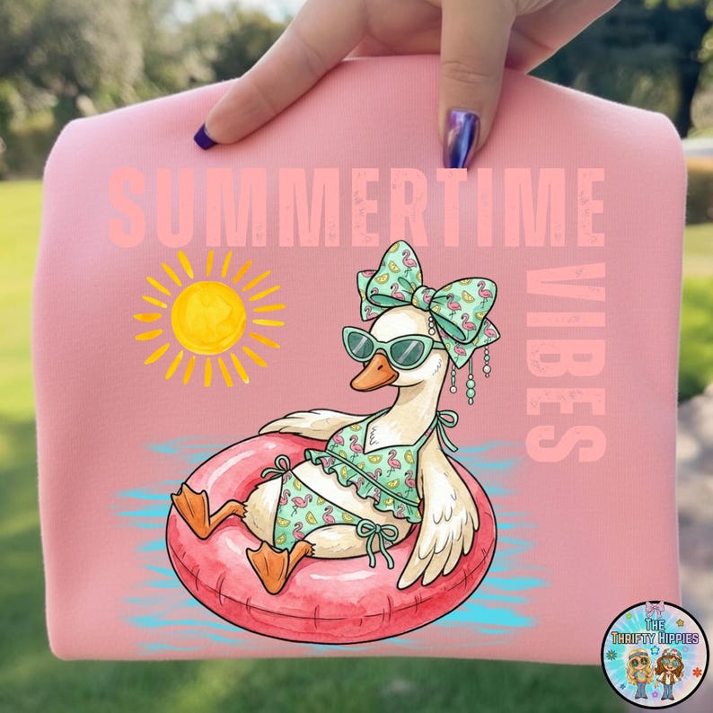 Puede incluir: Sudadera rosa con las palabras "SUMMERTIME VIBES" y un ganso de dibujos animados con gafas de sol y un traje de ba&ntilde;o, flotando en una c&aacute;mara de aire rosa. Un sol amarillo est&aacute; a la izquierda del ganso.