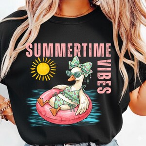 Puede incluir: Camiseta negra con un dise&ntilde;o de un ganso con gafas de sol, un lazo y un traje de ba&ntilde;o, flotando en un flotador rosa. Las palabras "SUMMERTIME VIBES" y un sol amarillo tambi&eacute;n est&aacute;n presentes.