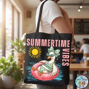 Puede incluir: Bolso tote negro con un gr&aacute;fico de un ganso con traje de ba&ntilde;o y gafas de sol flotando en una boya rosa. Las palabras "SUMMERTIME VIBES" est&aacute;n impresas en rosa y blanco. Tambi&eacute;n aparece un sol amarillo.