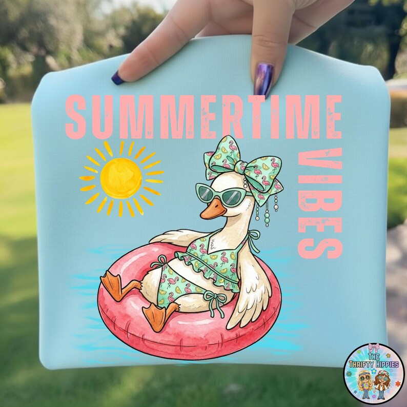 Puede incluir: Un art&iacute;culo azul claro con un dibujo de un ganso con traje de ba&ntilde;o y gafas de sol, flotando en un flotador rosa. Las palabras "SUMMERTIME VIBES" est&aacute;n impresas en rosa, con un sol amarillo. El art&iacute;culo es sostenido por una persona.