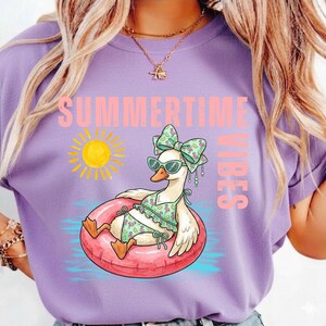 Puede incluir: Camiseta lila con la frase "SUMMERTIME VIBES" en rosa. El dise&ntilde;o presenta un ganso de dibujos animados con gafas de sol, un lazo y un traje de ba&ntilde;o, flotando en una boya rosa. Tambi&eacute;n se muestra un sol amarillo.