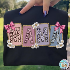 May include: Black sweatshirt with the word "MAMA" in leopard print letters. Each letter is in a different color, with a pink bow and daisies. The sweatshirt is held up by a hand.