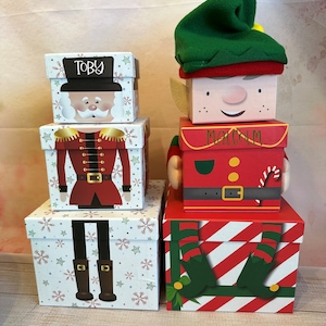 May include: Three red and white striped gift boxes with a Christmas elf design. The top box has the name "Malcolm" written on it. The middle box has a red and green elf design. The bottom box has the elf's legs and feet. Two smaller boxes are stacked on top of each other. The top box has a Santa Claus design and the name "Toby" written on it. The bottom box has a Nutcracker design.