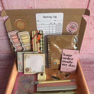 May include: A cardboard box containing a reading log, two book-shaped stickers, a floral bookmark, a spiral-bound notebook, a pink card with the text 'Your order made my day', a snail notepad, a small stapler, a resin ring, and several pens.