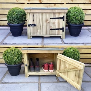 May include: A wooden shoe storage cabinet with a hinged door, black hinges, and a latch. The cabinet is made of light-coloured wood and has a small roof. Two potted plants with green foliage are on either side.