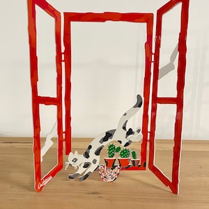 Puede incluir: Un marco de ventana de metal rojo con un gato blanco y negro saltando por la ventana. El gato corre hacia un cactus y tres plantas en maceta. El marco de la ventana está abierto y el gato está en el aire.