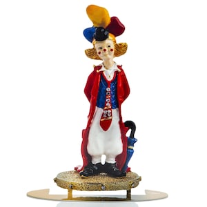 May include: A colorful clown figurine with a red, blue, and yellow hat, a white shirt, red tie, and red and blue jacket. The clown is holding a black umbrella and standing on a gold base with rhinestones.