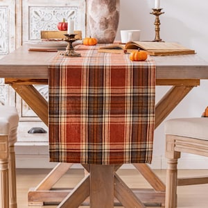 May include: A rectangular table runner featuring a plaid pattern in shades of burnt orange, cream, brown, and black. The fabric has a textured appearance and hangs over the edge of a wooden table, displaying a classic woven grid design throughout.