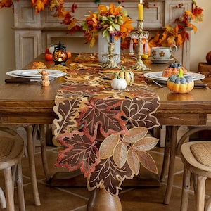 May include: A brown fabric table runner featuring intricate embroidered maple and oak leaf cutouts in shades of orange, red, and gold. The runner is displayed on a wooden dining table set with white plates, small decorative pumpkins, and candles.