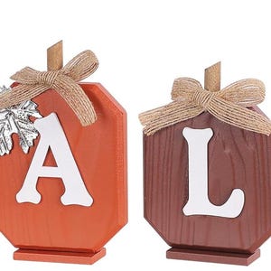 May include: Four wooden pumpkin-shaped decorations arranged to spell FALL. Each piece features a white letter, a burlap bow at the top, and a small wooden stem. The first and last are yellow, the second is orange, and the third is brown.