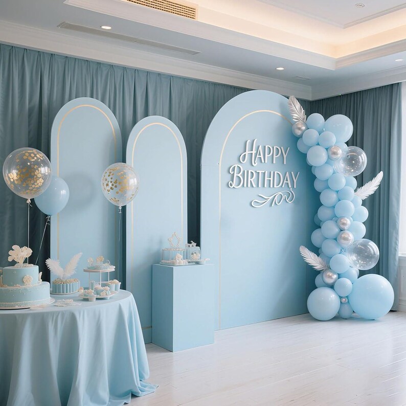 May include: A light blue birthday party backdrop with gold accents, balloons, and a table with a cake. The backdrop features the words "Happy Birthday" in white script. The scene is decorated with balloons and feathers.