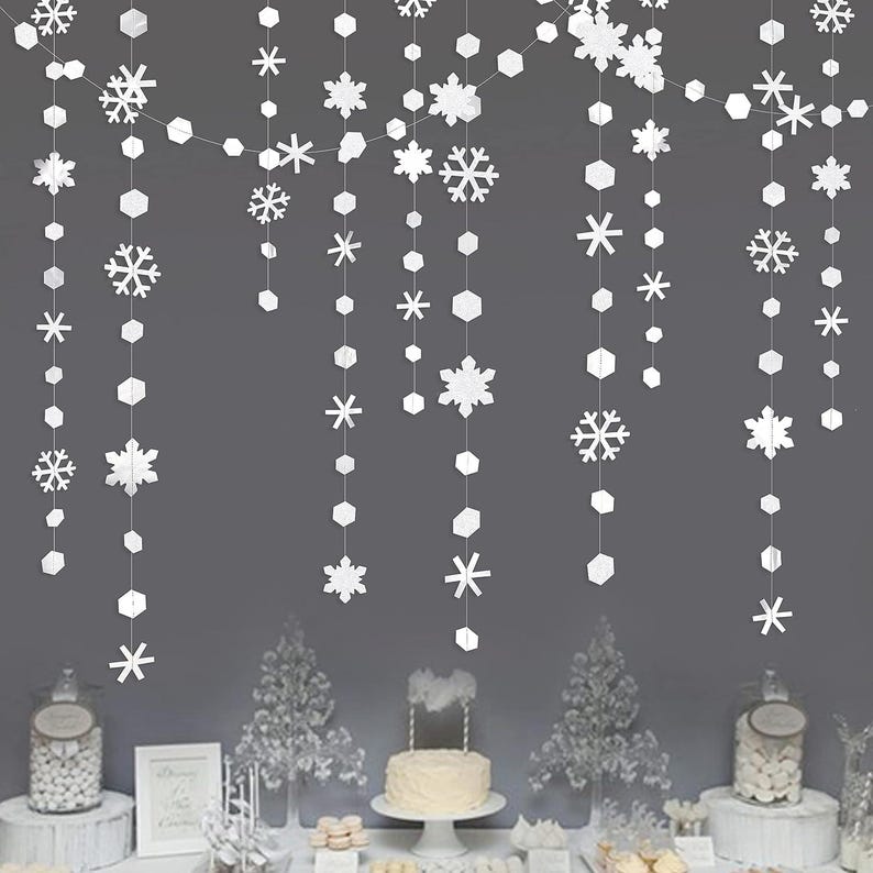 May include: White paper snowflake and geometric garland decorations hanging against a gray background. The garland features various snowflake designs and geometric shapes, creating a festive winter or holiday theme.