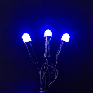May include: Three small, cylindrical light fixtures with black bases and white, dome-shaped tops that emit a bright blue glow. The fixtures are connected by black electrical wires that are coiled together at the bottom of the frame.