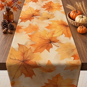 May include: A rectangular table runner featuring a repeating pattern of orange, yellow, and brown maple leaves on a cream-colored background. The fabric displays detailed leaf veins and varied autumn foliage shades across the entire surface area.