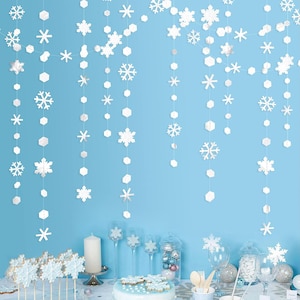 May include: White snowflake and geometric shape garlands hang against a light blue backdrop. The scene is set for a winter-themed party, with snowflake-shaped cookies on sticks, a cake, and decorative glassware.