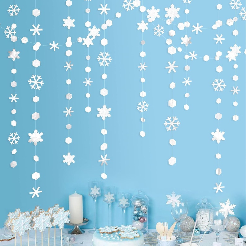 May include: White snowflake and geometric shape garlands hang against a light blue backdrop. The scene is set for a winter-themed party, with snowflake-shaped cookies on sticks, a cake, and decorative glassware.