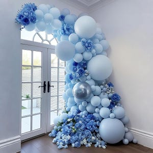 May include: A decorative balloon arch in shades of blue, including light blue and silver, adorns a doorway. The arrangement features various sizes of balloons and floral accents, creating a festive and elegant display for celebrations.