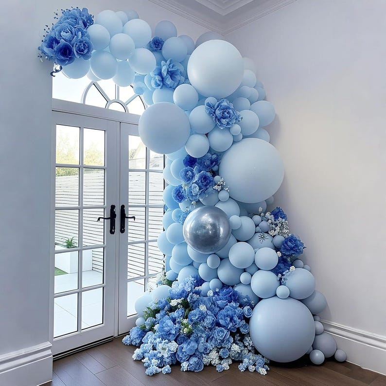 May include: A decorative balloon arch in shades of blue, including light blue and silver, adorns a doorway. The arrangement features various sizes of balloons and floral accents, creating a festive and elegant display for celebrations.
