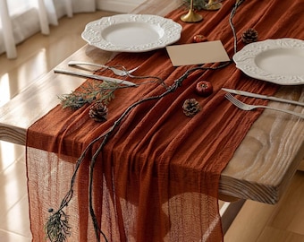 May include: A rust-coloured, crinkled fabric table runner draped over a wooden table. The setting includes two white embossed plates, silver forks and knives, a blank rectangular place card, small pinecones, and a decorative branch with berries.