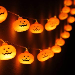 May include: A string of orange pumpkin-shaped LED lights, each featuring a carved jack-o'-lantern face with triangular eyes and a jagged mouth. The lights are connected by a thin wire and emit a warm, glowing light against a dark background.