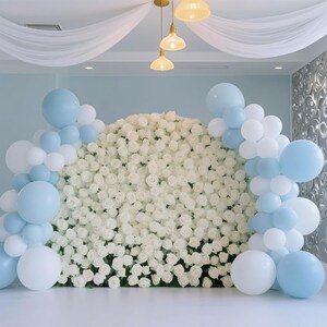May include: A backdrop featuring a wall of white roses, adorned with clusters of white and light blue balloons. The scene is illuminated by hanging lights and draped with white fabric, creating an elegant and celebratory atmosphere.