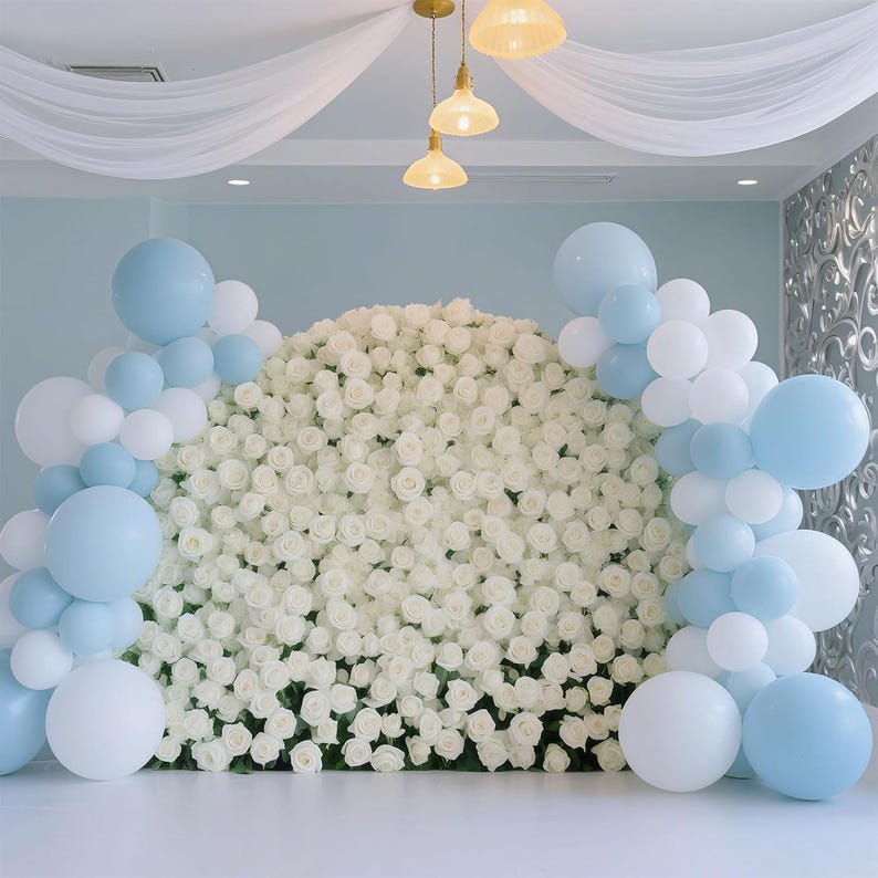 May include: A backdrop featuring a wall of white roses, adorned with clusters of white and light blue balloons. The scene is illuminated by hanging lights and draped with white fabric, creating an elegant and celebratory atmosphere.