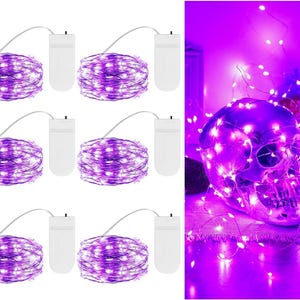 May include: Six sets of purple LED fairy lights on thin silver wire, each with a white battery pack. The lights are shown coiled and also wrapped around a human skull decoration, emitting a bright purple glow in a dark environment.