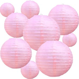 May include: A collection of light pink paper lanterns. The lanterns are round with a ribbed texture and a metal hook at the top for hanging. The lanterns are of varying sizes and are arranged against a white background.