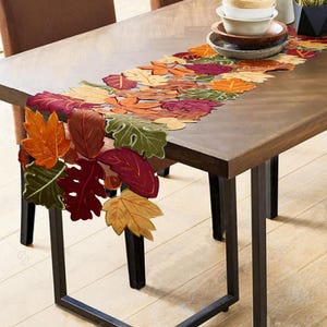May include: A table runner featuring a layered arrangement of fabric leaves in shades of orange, red, burgundy, and green. The runner is draped over a wooden table, with the leaf-shaped segments extending over the edge of the table surface.