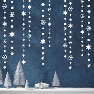 May include: White snowflake and geometric garland decorations hang against a dark blue background. Small silver Christmas trees and a candle holder with a lit candle are on the bottom. Festive holiday decor.