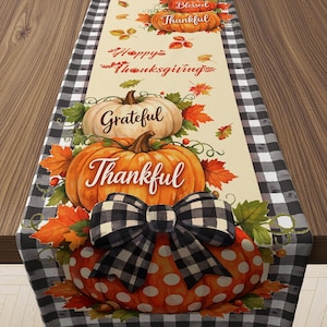May include: A table runner featuring a black and white buffalo check border and a cream center with pumpkins, autumn leaves, and text reading Blessed, Happy Thanksgiving, Grateful, and Thankful. A black and white checkered bow sits on a pumpkin.