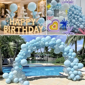 May include: A collection of party decorations, including light blue balloons, a "Happy Birthday" sign, and a balloon arch over a pool. The scene is set outdoors with palm trees and a fountain in the background.