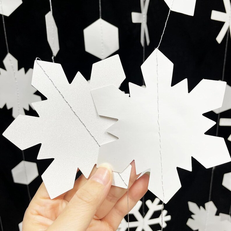 May include: White paper snowflake decorations hanging against a black background. The snowflakes are cut with intricate details and are connected by thin strings. Other geometric shapes are also visible.