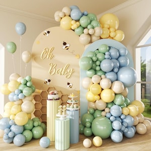 May include: A baby shower scene with pastel-colored balloons in blue, green, yellow, and cream. The backdrop features an arch with the words "Oh Baby" and bee decorations. There are cake stands and a cake on pedestals.