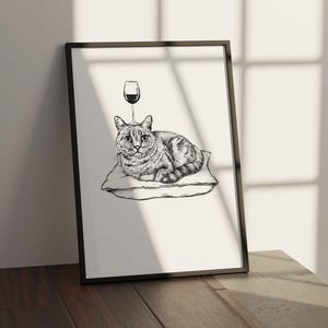May include: Black framed art print featuring a detailed pen and ink illustration of a cat resting on a pillow. A wine glass is drawn above the cat's head. The artwork is displayed on a wooden surface.