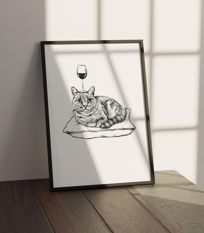 May include: Black framed art print featuring a detailed pen and ink illustration of a cat resting on a pillow. A wine glass is drawn above the cat's head. The artwork is displayed on a wooden surface.