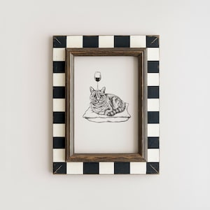 May include: A framed black and white illustration of a cat resting on a pillow with a wine glass above its head. The frame has a black and white checkered pattern. The artwork is displayed on a white wall.