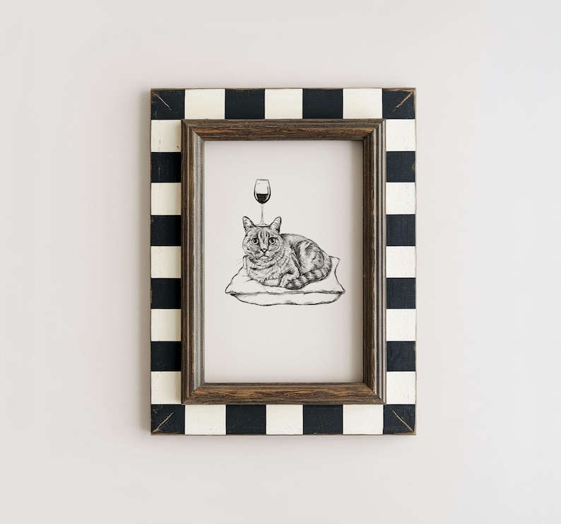 May include: A framed black and white illustration of a cat resting on a pillow with a wine glass above its head. The frame has a black and white checkered pattern. The artwork is displayed on a white wall.