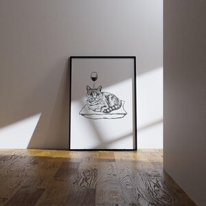 May include: Black and white framed art print featuring a detailed illustration of a cat resting on a pillow. A wine glass is drawn above the cat's head. The artwork is displayed on a wooden floor.