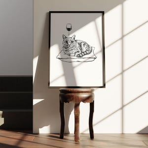 May include: Black framed art print featuring a cat resting on a pillow with a wine glass above its head. The cat is depicted in a detailed black and white illustration. The print is displayed on a dark wooden side table.