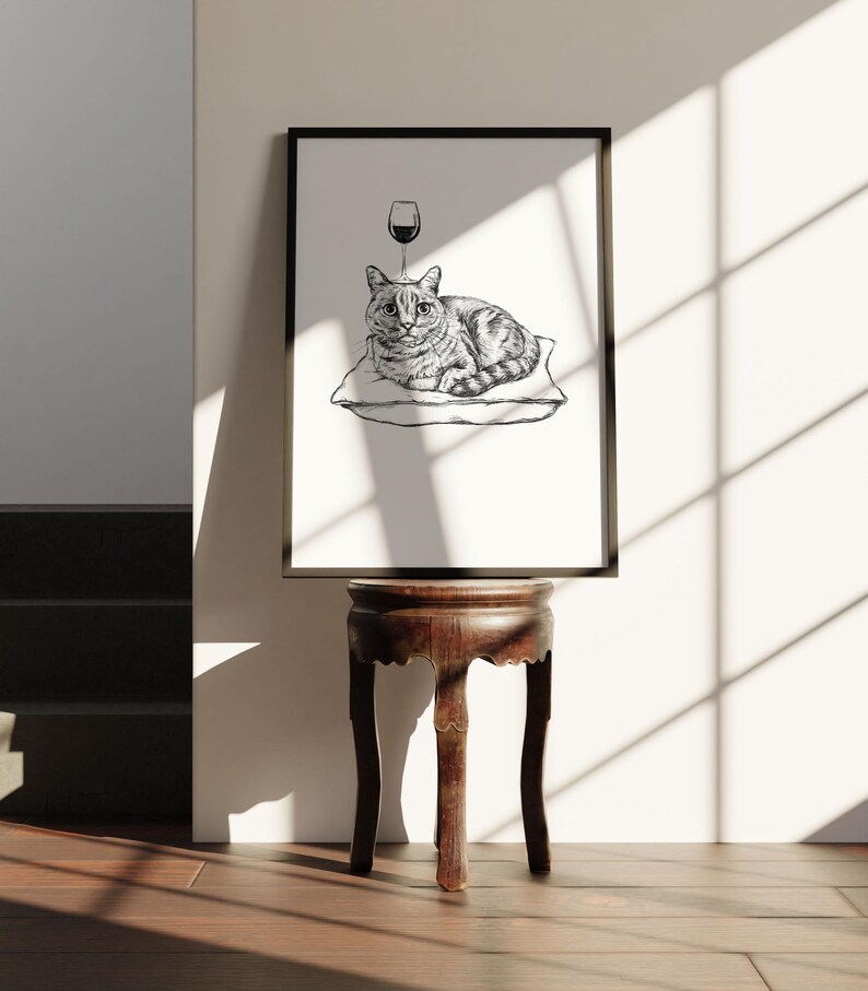 May include: Black framed art print featuring a cat resting on a pillow with a wine glass above its head. The cat is depicted in a detailed black and white illustration. The print is displayed on a dark wooden side table.