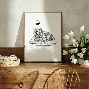 May include: A framed black and white illustration of a cat lying on a pillow with a wine glass above its head. The artwork is displayed on a wooden dresser with a woven basket and a vase of white tulips.