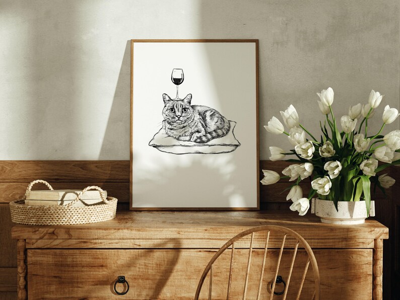 May include: A framed black and white illustration of a cat lying on a pillow with a wine glass above its head. The artwork is displayed on a wooden dresser with a woven basket and a vase of white tulips.