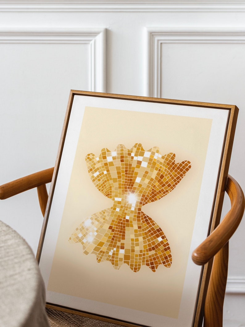May include: A framed print of a gold bow tie pasta shape made of small squares, resembling sequins. The pasta is on a light beige background.