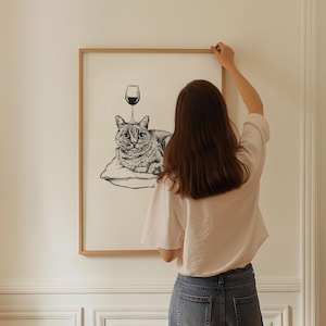May include: Black and white illustration of a cat lying on a pillow with a wine glass above it. The artwork is framed in a light brown wooden frame. The image is hanging on a white wall.