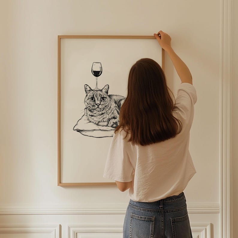 May include: Black and white illustration of a cat lying on a pillow with a wine glass above it. The artwork is framed in a light brown wooden frame. The image is hanging on a white wall.