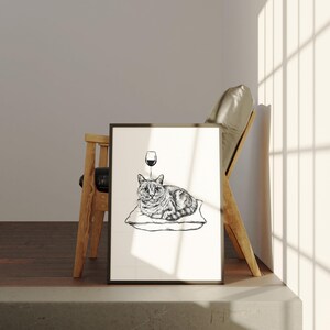 May include: Black and white framed art print featuring a cat lying on a pillow with a wine glass above its head. The cat has striped markings. The art print is displayed next to a wooden chair.