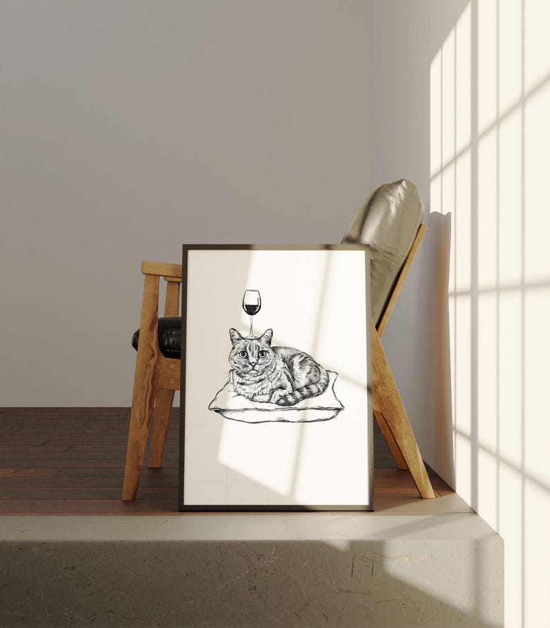 May include: Black and white framed art print featuring a cat lying on a pillow with a wine glass above its head. The cat has striped markings. The art print is displayed next to a wooden chair.