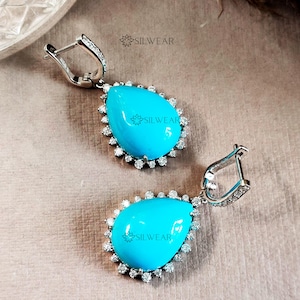 May include: A pair of silver teardrop earrings with turquoise stones set in a halo of clear stones. The earrings have a hook fastening.