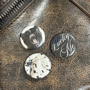 Op de afbeelding: Drie ronde buttons. Eén button toont een gapende zwarte kat, een andere is wit met zwarte stippen en de derde is zwart met de witte cursieve tekst Lucky Me. De buttons hebben een glanzende afwerking en zijn in een driehoekig patroon gerangschikt.